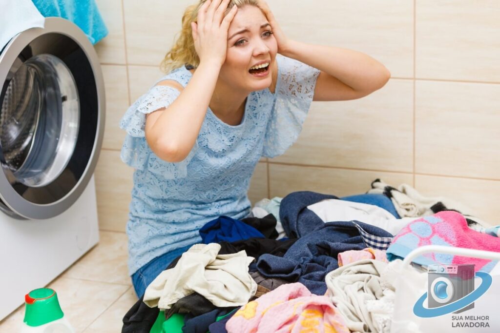 Mulher com expressão de frustração sentada no chão, com a cabeça nas mãos, ao lado de uma pilha de roupas sujas e uma máquina de lavar roupa. A imagem ilustra o problema de roupas que não lavam direito.