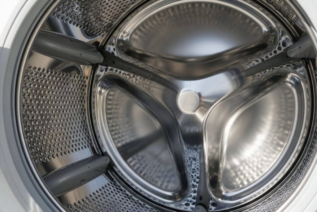 Visão interna de um tambor de aço inox de máquina de lavar, completamente limpo e brilhante, refletindo a luz.